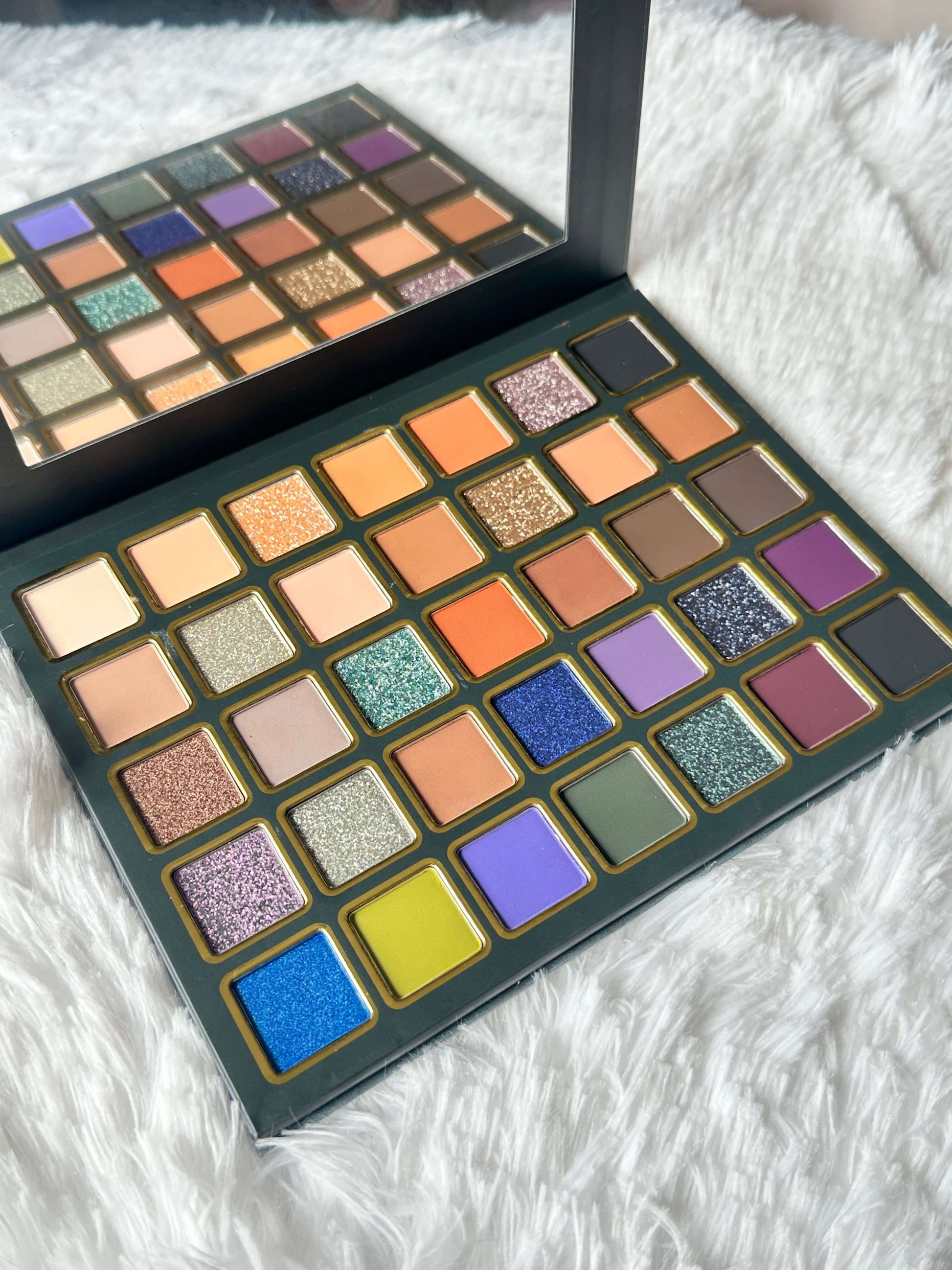A large eyeshadow palette with multiple square pans of colorful makeup, shown open on a white faux-fur surface. The palette has shades including beige, orange, brown, green, blue, purple, and pink, with a mirror visible inside the upper section.