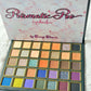 An open eyeshadow palette from Cessy Charm Cosmetics,  featuring the 'Prismatic Pro' shade name on the inner lid. The palette contains 24 square eyeshadow pans in a variety of colors including gold, orange, blue, purple, green, and pink, set in a dark teal case with gold trim, resting on a white textured fabric background.