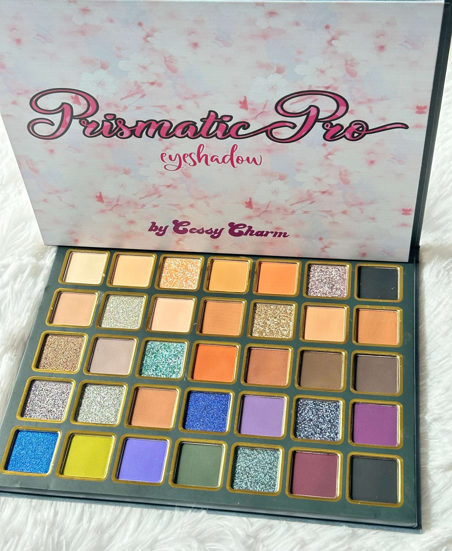 An open eyeshadow palette from Cessy Charm Cosmetics,  featuring the 'Prismatic Pro' shade name on the inner lid. The palette contains 24 square eyeshadow pans in a variety of colors including gold, orange, blue, purple, green, and pink, set in a dark teal case with gold trim, resting on a white textured fabric background.