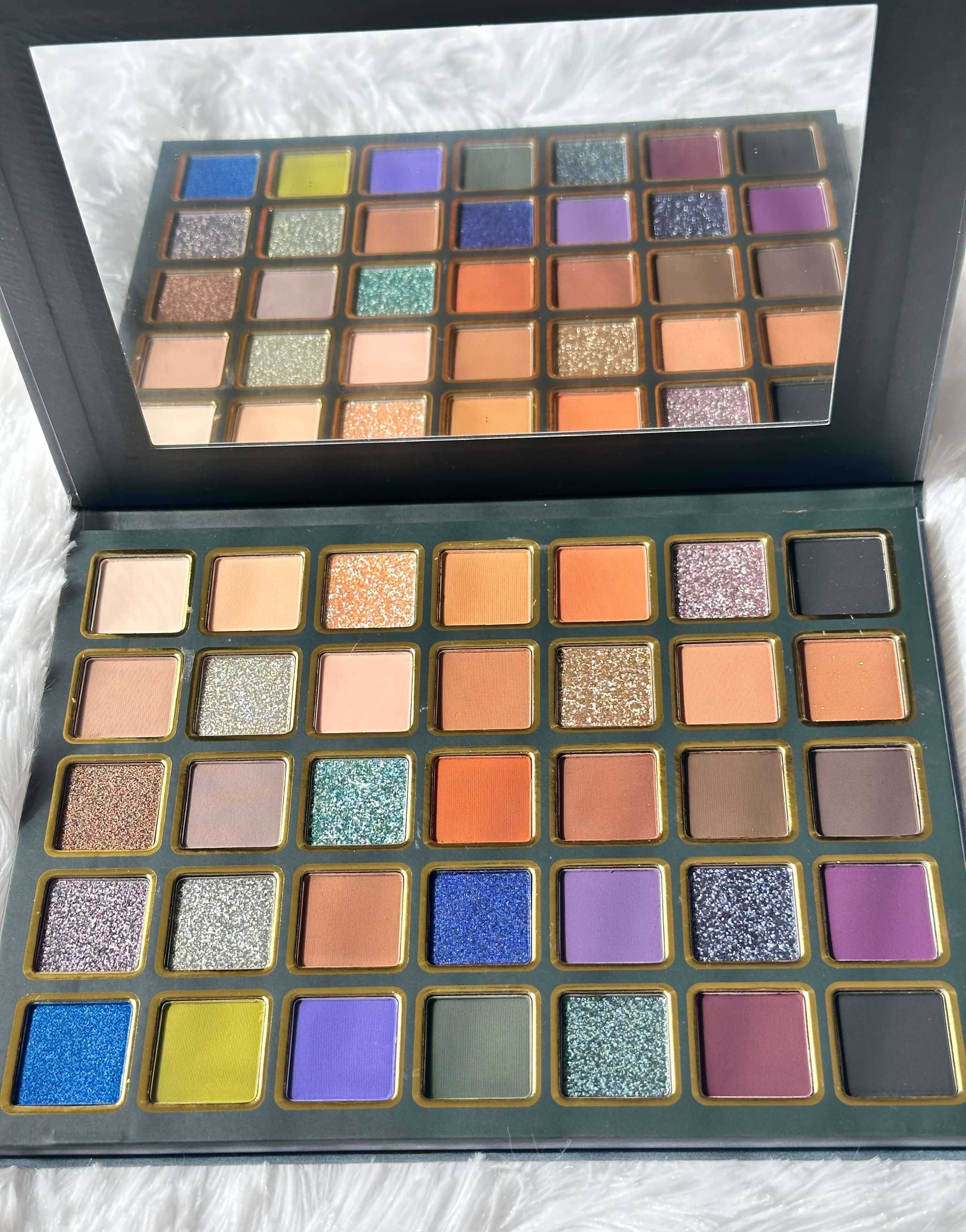 An open eyeshadow palette from Cessy Charm Cosmetics,  featuring the 'Prismatic Pro' shade name on the inner lid. The palette contains 24 square eyeshadow pans in a variety of colors including gold, orange, blue, purple, green, and pink, set in a dark teal case with gold trim, resting on a white textured fabric background.